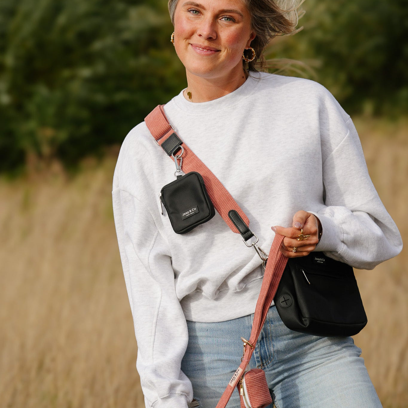 Vrouw houdt de koraalkleurige corduroy riem uit de Jaap & Co Golden Trails collectie vast, inclusief poepzakjeshouder, in warm nazomerlicht.