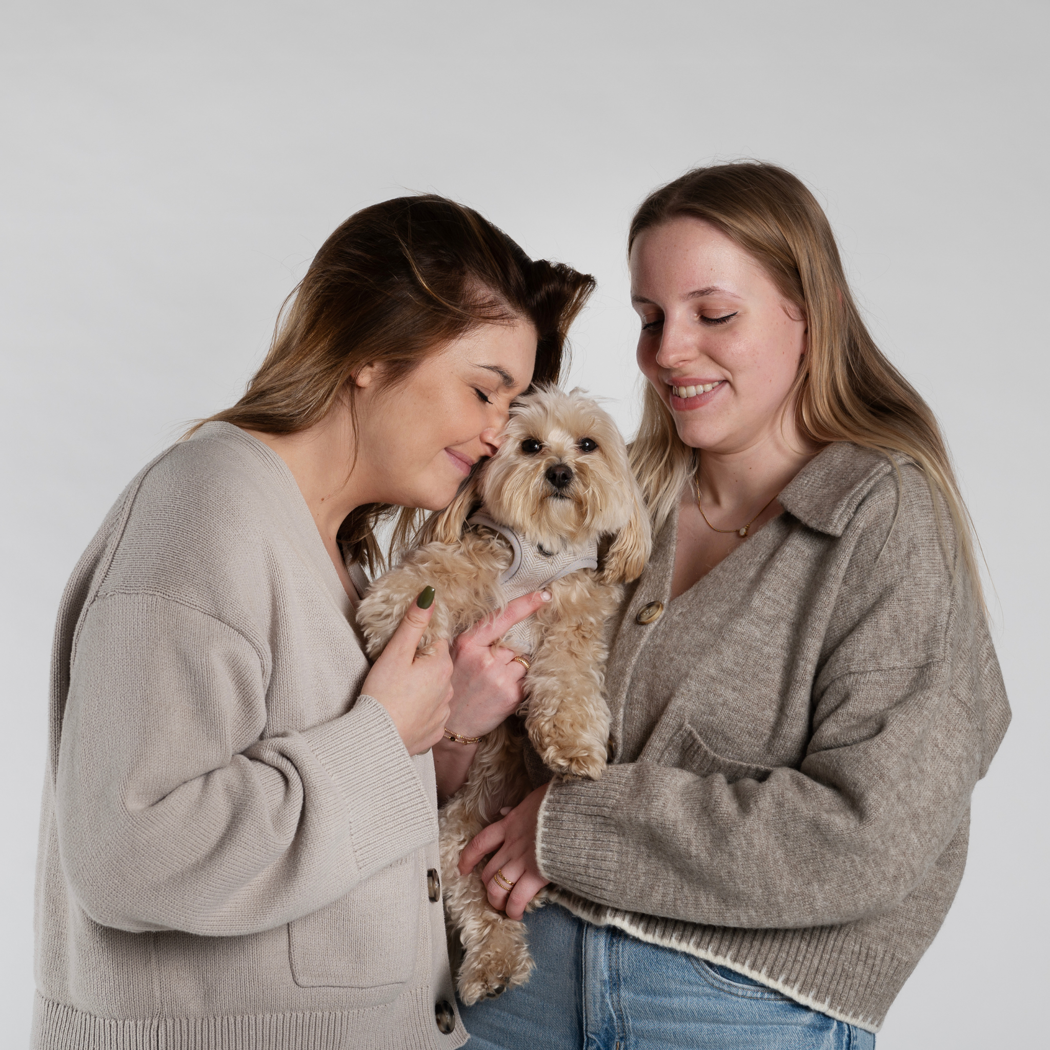 Twee modellen knuffelen een maltipoo hondje dat het Jaap & Co hondentuigje uit de Classic Beige collectie draagt – liefdevol en stijlvol moment.